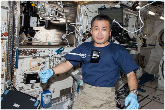 In front of the Fluorescence Microscope (upper left) installed on Kibo, Astronaut Wakata poses for a photo before setting the sample chamber (floating below his face).